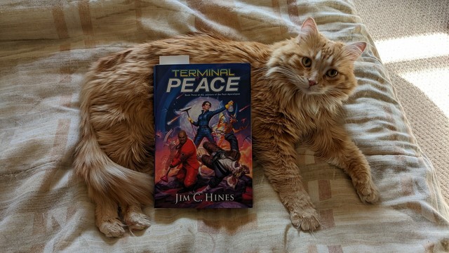 The book Terminal Peace, by author Jim C. Hines, laying on a fluffy orange cat, who is resting on a comforter on a bed.