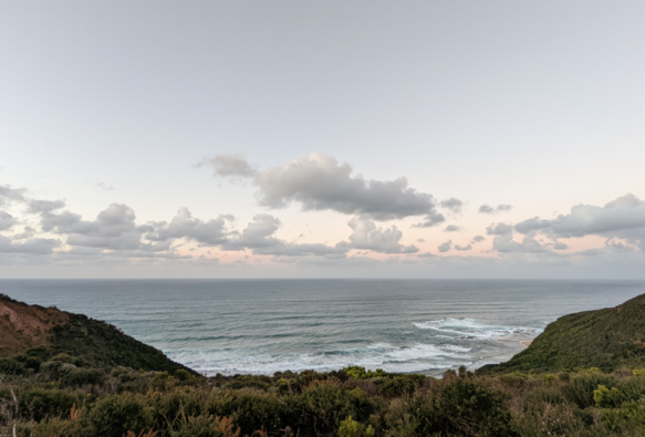 Sunset view from the cliff-top toilet near Devil's Kitchen Campground, Great Ocean Walk, Victoria, Australia. In the foreground, windswept coastal scrub clings to rocks and steeply descending sand-dunes. In the mid-ground is a blue-green sea with rows of gentle white-capped waves. The sky fades from blue at the top to peach-pink in the middle with puffy grey clouds that merge into a hazy horizon.