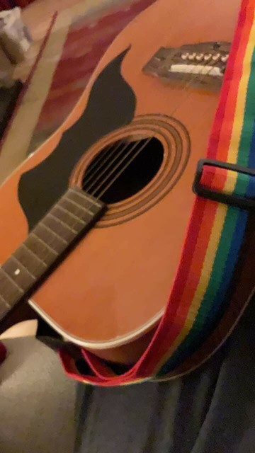 close up slightly blurred photo of a guitar body and a few inches of neck. A rainbow strap draped across the bottom.