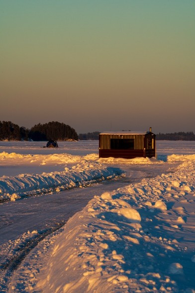 A permanent ice-fishing shelter sits at the end of a plowed “road” on White Iron Lake, MN as the sun says.  A smaller temporary shelter can be seen in the background.