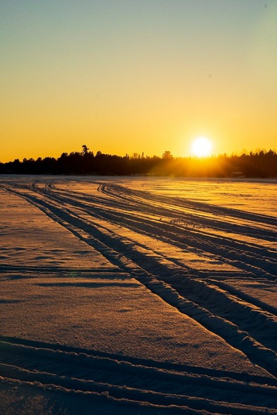 The sun sets behind trees over a snowy snowmobile-tracked lake.
