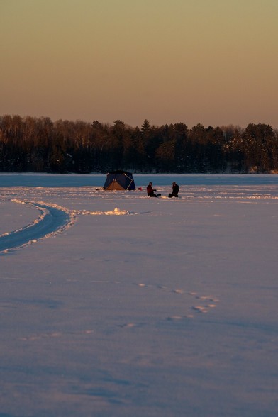 Two humans sit outside a temporary ice fishing shelter on a frozen lake illuminated by the setting sun.