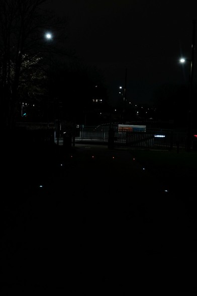 White and red lights create a rectangular area in an otherwise dark area of ground. There are lights above them.There's a barrier and a slightly better lit road, then the lights begin again, curving to one side, with streetlights above.