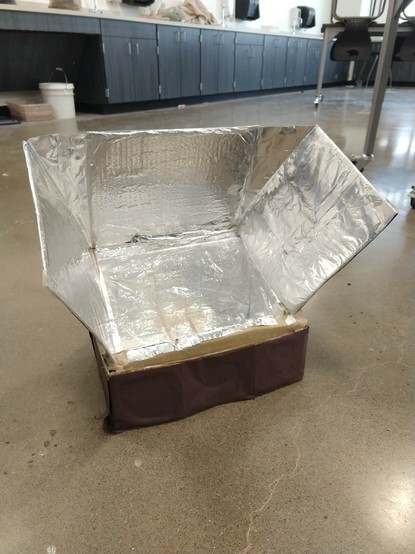 Picture of solar box oven made from small cardboard box, with aluminum foil reflective panels, clear window in slanted lid.