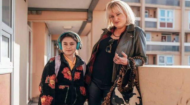 Daisy May Cooper stands beside her daughter, who is wearing headphones, in an office in Rain Dogs