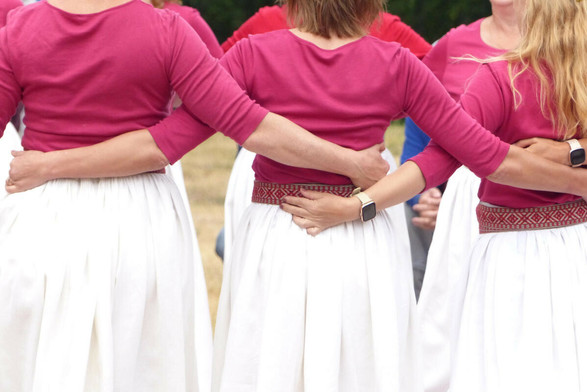 trois danseuses, de dos, se tiennent par la taille. Elles ont les mêmes T-shirts, jupes et ceintures, évoquant un costume traditionnel mais pas trop. Des montres informatisées sont visibles sur deux poignets.