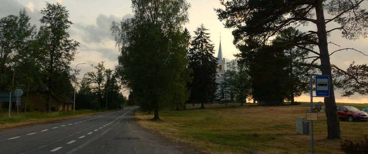 La "rue" principale du village : une route bordée de de pelouse, d'arbres et de lampadaires LED. Sur le côté droit, le panneau d'un arrêt de bus et quelques voitures garées sur un parking d'herbe. On aperçoit aussi une maison de bois et l'église au fond, entre de grands épicéas.