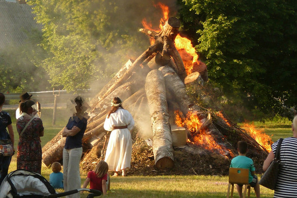 Des troncs, empilés en pyramides, commencent à brûler devant des gens qui discutent. Une jeune femme de dos, en robe de lin blanc, fleurs dans les cheveux, vient de mettre le feu.