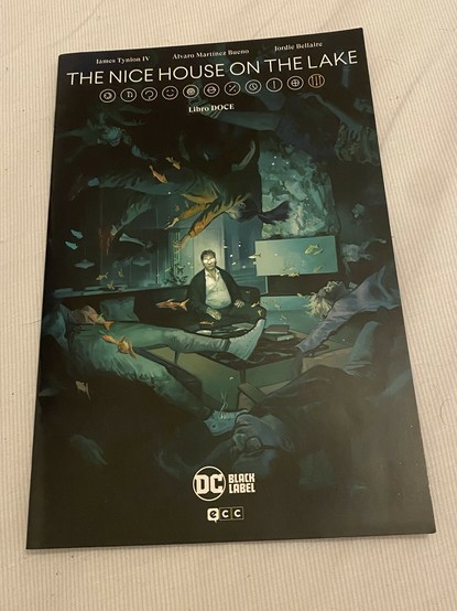 Una copia del número 12 de The nice house on the lake apoyada sobre la cama. La portada es muy surrealista y me va a costar describirla, pero ahí va mi intento. En en salón de una casa rústica se ve en el foco a un hombre desaliñado sentado en un sofá. Lleva una camisa blanca algo desabotonada y sus gafas reflejan mucho la luz. Da la impresión casi de que sus ojos iluminan. La estancia parece estar sumergida porque se ven peces y el resto de las personas que hay en el salón flotan inconscientes en posiciones extrañas.
