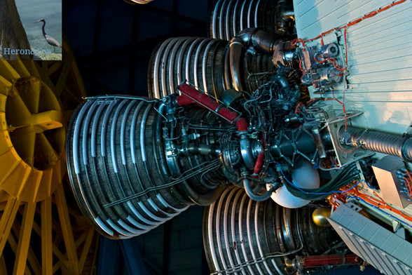 An indoor photograph of five rocket metal rocket engines, with many complicated hoses and pipes and wires, attached to a white metal cylinder. The engines are the five Jay Two rocket engines that powered the S Two upper stage for the Saturn Five rocket, which took humans to the Moon.