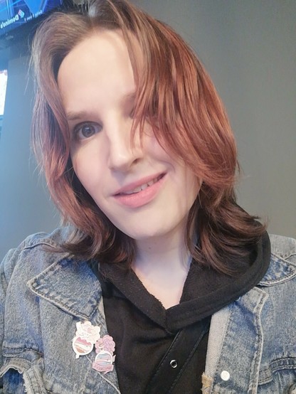 Selfie von Lili vor einer grauen Wand. Sie lächelt leicht. Das Haar liegt ungekämmt halb im Gesicht, oben zum Mittelscheitel mit leicht Volumen im Pony. Man sieht nur etwas grauen Lidschatten auf den Augen, sonst komplett ungeschminkt.

Sie trägt eine Jeansjacke und darunter einen schwarzen Hoodie.