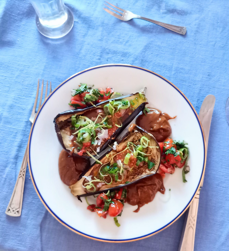 Auf dem Teller zwei halbe gegrillte Auberginen, darauf, Frühlingszwiebeln, Tomatenstückchen, und eine braune Creme