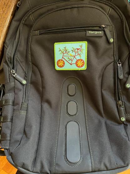 The same black backpack now has a charming rectangular patch depicting a stylized bicycle made from brown branches and green leaves. The wheels have a bright yellow sunflower centered in a brown background. It is infinitely more delightful than it was previously.