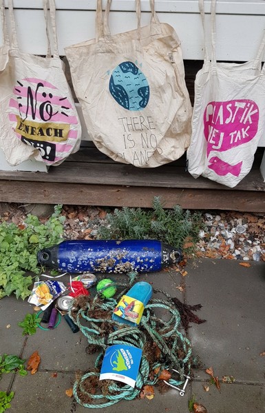 Gesammelter Strandmüll.
Im Hintergrund 3 waschbare Stoffbeutel, in denen ich den Müll sammle, den ich tragen kann.
 Im Vordergrund ein PlastikfreeHindsholm-Aufkleber.

Auf dem ersten Stoffbeutel der Aufdruck: Nö Einfach Nö!
2ter Aufdruck: There is no planet B
3ter Aufdruck: Plastik ne tak