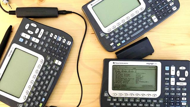 Three Texas Instruments Voyage 200 graphing calculators on a wooden surface