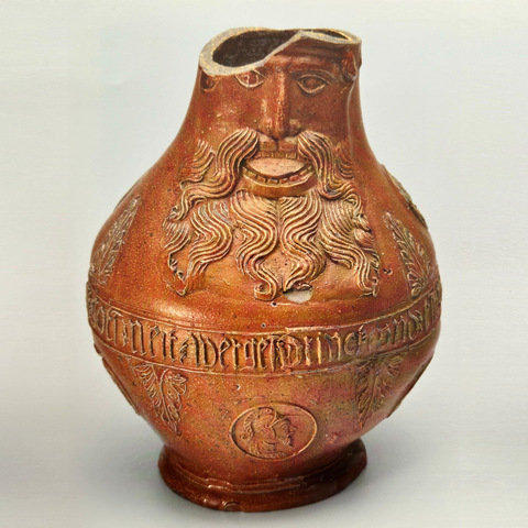 On this image is a sixteenth century Bellarmine jug, which features relief decoration of a bearmed man sticking out his tongue. The brownish jug is made from stoneware, salt glaze and engobe, and was acquired in 1990. The top of the jug has some damage. Around the belly of the jug runs a text band which states in Gothic letters: 'Drink und est Gott neit wergest' (Eat and drink but forget not god).