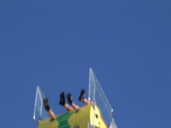 Am unteren Rand des Bildes schwingt eine gelbe Gondel eines Jahrmarkt-Fahrgeschäftes in den blauen Himmel. Vier Kinderbeine strecken sich aus der Gondel in den Himmel. Das Bild ist leicht verwischt (Bewegungsunschärfe).