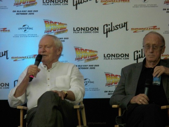 Julian Glover + Ken Colley sitting while on stage at London Film & Comic Con, talking about when they were both in Star Wars, for one. Two old men probably in their mid-seventies.

Glover looks animated while telling some tale from back in the day. He has his left hand up on his left knee.

Colley is looking almost at us, but his eyes are a bit more to his left. He has the mic down in his right hand and holds a small bottle of water closer to his chest on his left side.