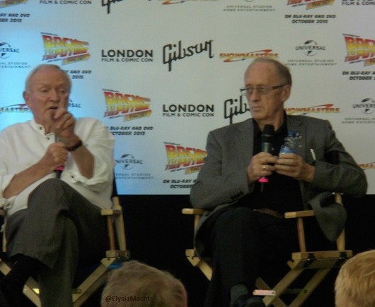 Same setting as the first picture, a moment earlier.

Glover to our left has a white shirt + grey trousers; and talking into his mic, holds a finger up while making some point. Colley has glasses and is wearing black other than his grey blazer. He's holding both the mic and the small water bottle up, while looking at something in the distance & out of our sight, to his right.