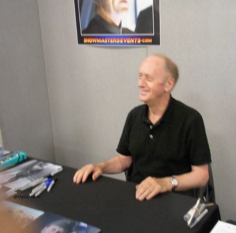 Ken Colley signing at London Film & Comic Con, seated behind a table with pens and promo pics in front of him, to choose what we want signed. It was hot so he has taken off the jacket.

He's smiling while looking at whichever person was there before me, not seen.