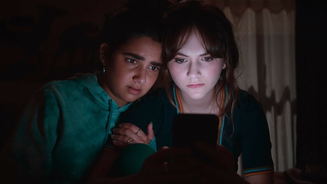 Geraldine Viswanathan schaut Emilia Jones besorgt über die Schulter, Emilia schaut auf ihr Handy. Beide sitzen in einem dunklen Raum, das Handydisplay beleuchtet ihre Gesichter.
