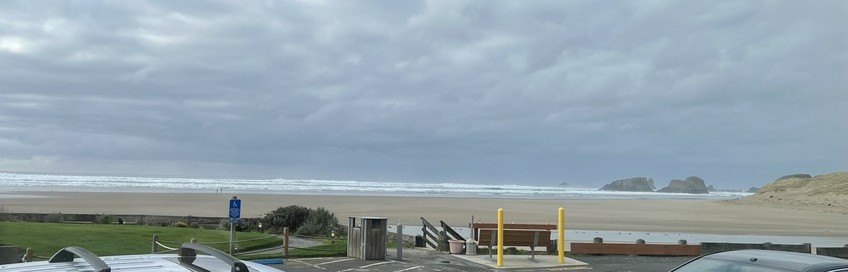 Low tide at Cannon Beach, Oregon- a broad expanse of sand with Ecola Creek flowing through it to the sea, with a tan sandy headland and rocky spires on the left fading to the distance. The old Tillamook lighthouse is barely visible beyond the sea spires. The sky is cloudy, the sea is white-capped.