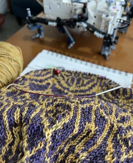 A hand knit sweater in purple and old rests on a paper spiral-bound pad, with a partial ball of yellow gold next to it. The legs of a medium-sized  Lego project are blurry at the top.