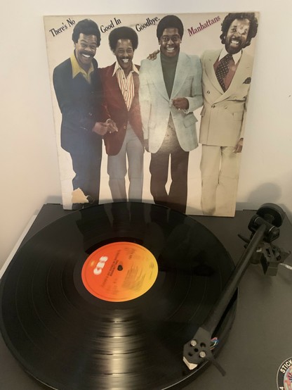Ein Foto einer Schallplatte, die auf einem Plattenspieler läuft, mit der Plattenhülle im Hintergrund, auf der die Gruppe "Manhattans" abgebildet ist, vom Album „There‘s no good in goodbye“.
