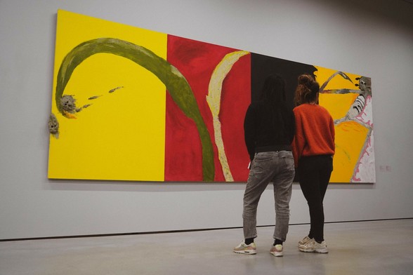 Two women stand before a large painting by Italian artist Mimmo Paladino. The painting consists of four large colour blocks next to each other: yellow, red, black and orange. On the yellow block, there are two protruding elements, which resemble faces. The red block features two organic shapes, one of which also overlaps the yellow block. The orange block contains a human-like figure.