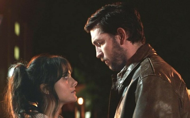 Emilia Jones and Nicholas Braun on a dark street. They are looking in each others eyes and standing fairly close together.