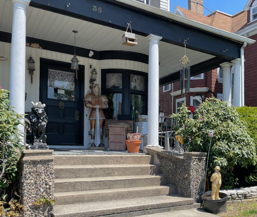We are looking at the front porch of a house. Concrete steps up to the porch are flanked by short stone pillars. In front of the right pillar is a golden figure of a firefighter, holding an ax propped up in a tub for a potted plant.  Atop the pillar on the left is a stone lion painted black with a silver-painted crown. On the porch to the right of the door is a suit of armor in shades of copper, silver, and gold. To the right of the armor beneath the bay window is a short rattan chest of drawers, next to that several plant pot stacked in each each other, then a jumble of other objects that we can't see clearly. Hanging from the overhang of the porch is a small bird feeder and a set of wind chimes. In the window on the door and in the panes of the bay window are lace curtains. a hanging lantern-style light hangs in front of the door.  Video recorded by a camera above the door must be entertaining.