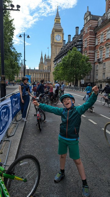 8 year old boy (dressed in green next to his green bike) stood in front of Houses of Parliament, London. Other cyclists fill the background.