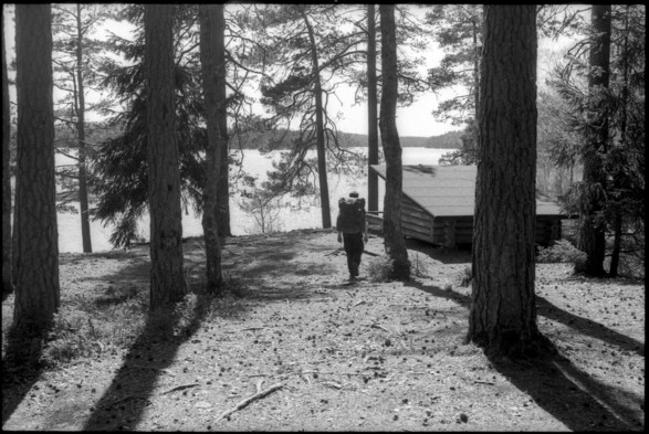 A black and white photograph of a hiker approaching a shelter by a lake. The sun is shining, making the pine-trees casting sharp shadows on the ground. Between the trees, a large lake can be seen.