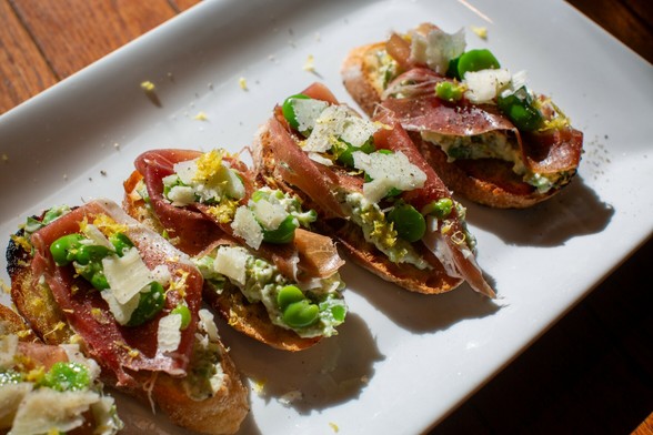 Photograph of several grilled sections of bread topped with green purée, thinly-slide dried meat, shreds of a hard cheese, dark-green beans, lemon zest, salt, and pepper.