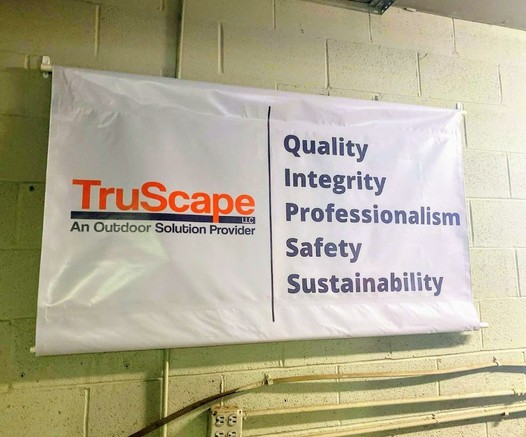 A banner hanging on a wall with the TruScape LLC logo and the tagline "An Outdoor Solution Provider." The banner lists the company's core values: Quality, Integrity, Professionalism, Safety, and Sustainability.