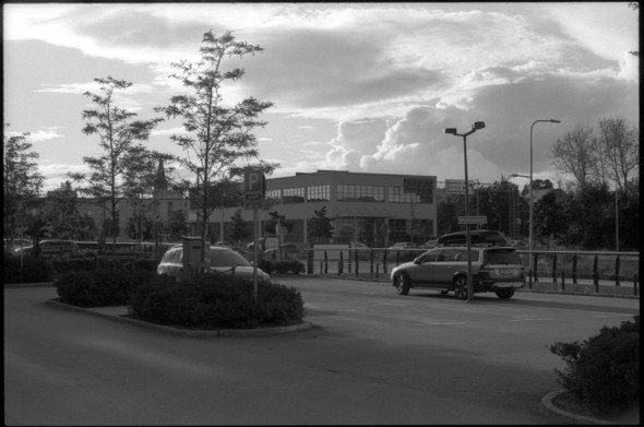 A black and white photo taken from a parking lot. There are a few cars parked there, and some buildings can be seen in the distance, along with a church tower. The clouds in the sky above the scene is very dramatic and it seems that there is rain coming in soon.