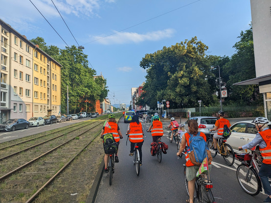 Auf einer Fahrbahn fährt eine Gruppe von Radfahrenden unterschiedlichen Alters. Die Gruppe wird von einem Fahrzeug der Polizei angeführt.