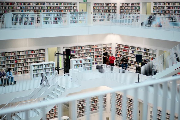 Eine große Halle, komplett in Weiß, mit mehreren umlaufenden Galerien. Entlang der Wände stehen überall Bücherregal. In der gegenüberliegenden Ecke tritt ein Jazz-Trio mit Kontrabass und Gitarre auf. Die Sängerin trägt eine intensiv-rote Jacke und bildet den Mittelpunkt des Bildes.