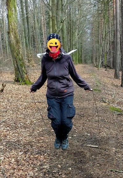 Frau beim Springseil springen im Wald. Die Aufnahme ist aus dem Frühjahr. Die Bäume haben keine Blätter und Laub liegt auf dem Waldboden. Das Gesicht ist mit einem lustigen Smilie verdeckt.