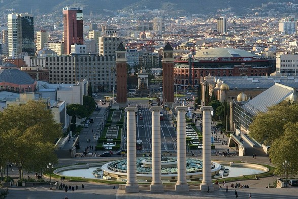 Une place en Espagne bordée d'immeubles et de quatre pylônes en pierre au premier plan. Au fond un enchevêtrement de maison, immeubles et bâtiments.