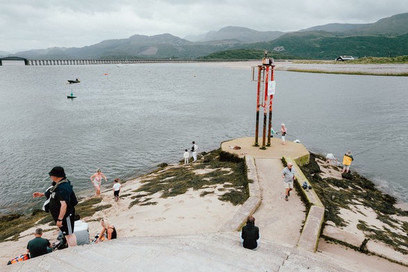 Various small groups of people are fishing, crabbing and enjoying the view at the end of a tide wall. In the distance there are mountains.