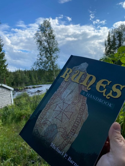 Photo of a book being held up in the foreground. The title of the boom is “Runes”, with a smaller subtitle below it “A Handbook”. A runestone takes up most of the cover of the book. In the background of the photo is a stream, a few birch trees, the edge of a small log cabin to the left, and a pine forest on the far side of the stream. Above is a blue sky with a few white clouds.