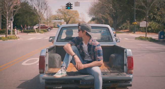 A teen boy sits casually on the back of an open pickup truck bed as it drives down the street