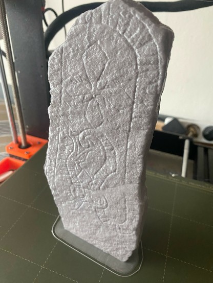 Photo of a silvery gray plastic runestone standing on the print surface of a 3D printer. It’s possible to see some of the ornamentation on the stone. At the top is a cross, below it is the head of a snake eating its own tail. The snake continues around the edge of the stone and contains barely visible runes.