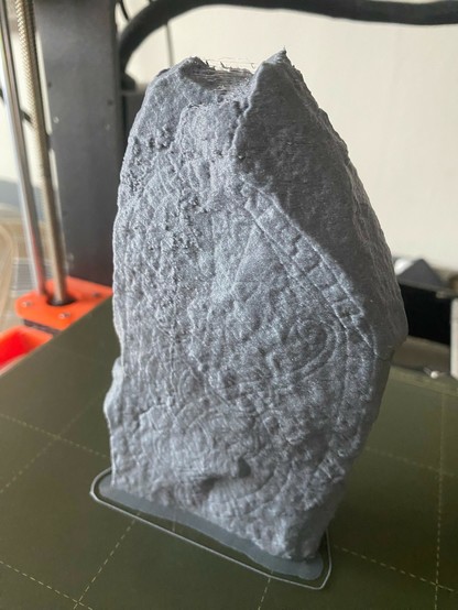 Photo of a 3D printed runestone model standing on the bed of a 3D printer. It’s gray, with some layer lines showing. It’s possible to make out some of the carved ornamentation, but it is very faint.