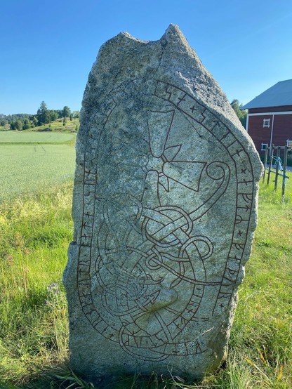 Photo of the runestone in a field of green grass. The stone is gray, with painted flowery red ornamentation. A rune band goes around the edges and crosses at the bottom. A large cross is carved in the top of the stone. In the background to right is a red barn with white corners.