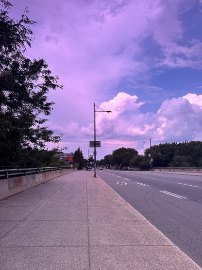 A wide sidewalk and a road stretch into the distance under a partly cloudy sky with shades of purple and pink, seen through sunglasses. On the left, a row of trees lines the path, and a McDonald's sign is visible in the background. The sky's color shifts between lavender and blue, giving the scene a surreal, tinted appearance.