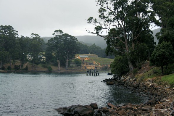 A narrow harbour, with rocky shores and tree-covered banks. 
The foreground is a spit of rocks curving out into the grey water. On the bank are gum trees and green bushes, almost leaning over the water.
Just behind is a wooden jetty.
In the background, slightly misty, the land is cleared and are a scattering of yellow brick buildings, and a small, orange cottage. There's a house visible amongst the trees on the far bank.
