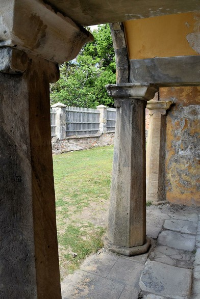 The lower level of the tower, from inside. A row of columns, holding up arches, painted yellow on the inside. The arches to be stone, narrowing at the top and then a wide support. 
Beyond the arches, is a stretch of lawn, worn in places, and bordered by a wooden fence with stone pillars, and green leafy trees beyond.