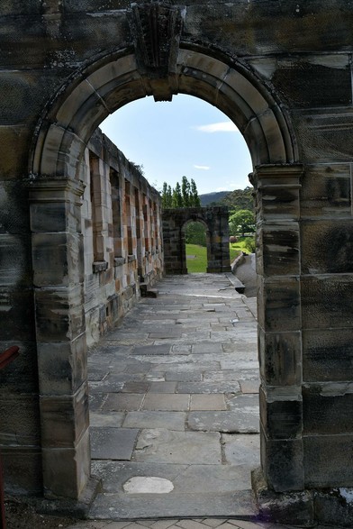 Looking through a stone arch, along a walkway paved with stone pavers to another arch. Alongside the walkway is a wall, with a row of narrow windows, looking like the front of a building. It was once, but now, after a series of fires, it's just walls and empty windows.
Beyond the far arch, trees and lawn can be seen